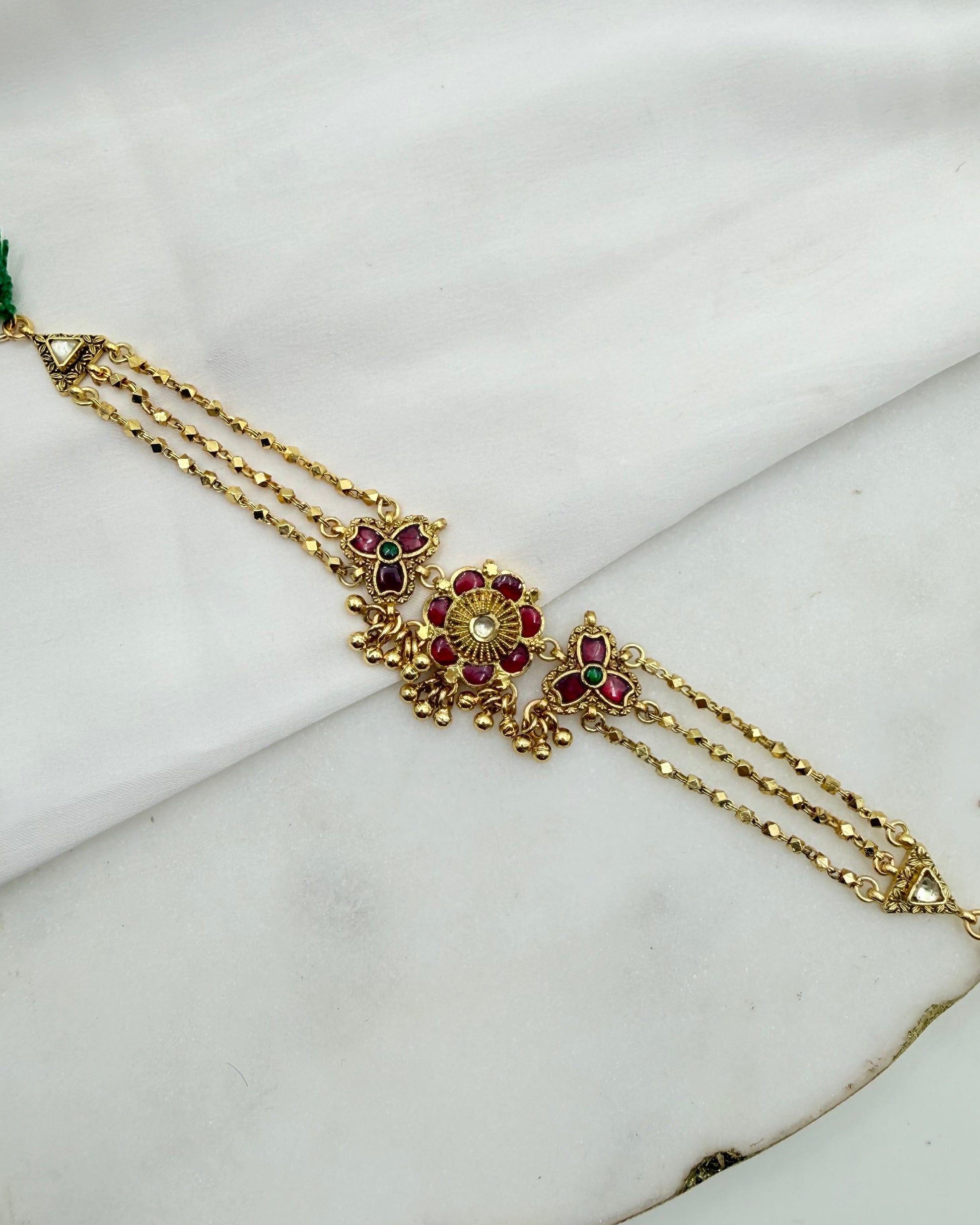 Gold-toned traditional Indian jewelry piece featuring a central floral motif with red and green stones, displayed on a white marble surface.