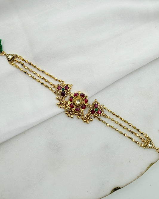 Gold-toned traditional Indian jewelry piece featuring a central floral motif with red and green stones, displayed on a white marble surface.