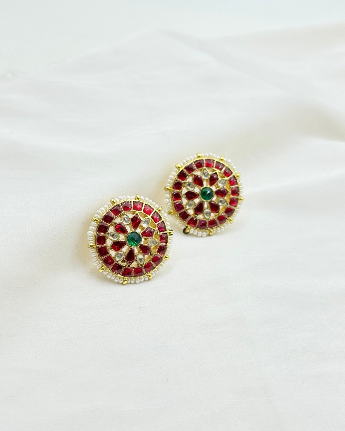 A pair of gold-plated Kundan earrings featuring red stones, a central green stone, and white stones, displayed on a soft white fabric background.