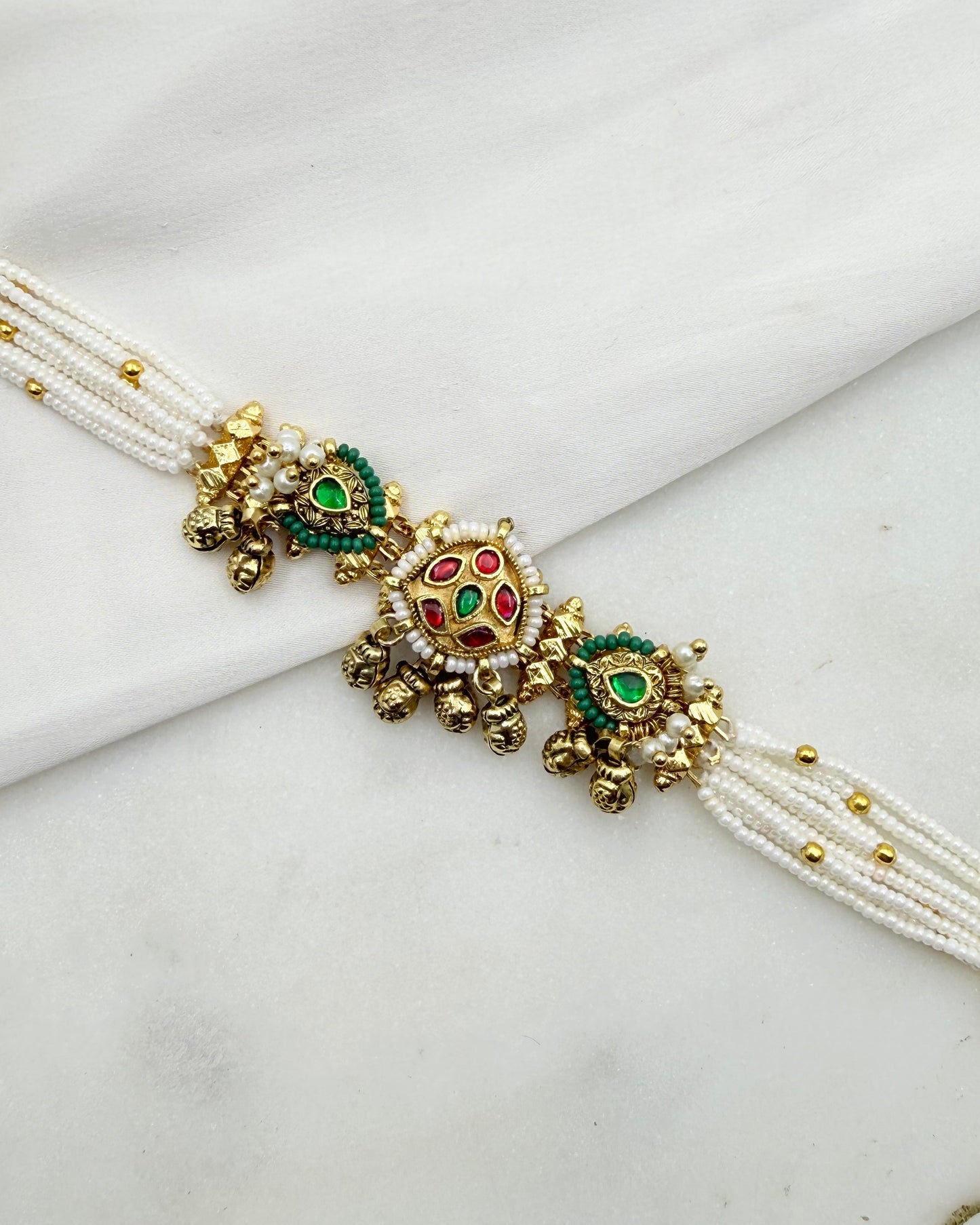 A white beaded wristband or rakhi with ornate gold-toned decorative elements, featuring green and red enamel details, laid on a white fabric background.