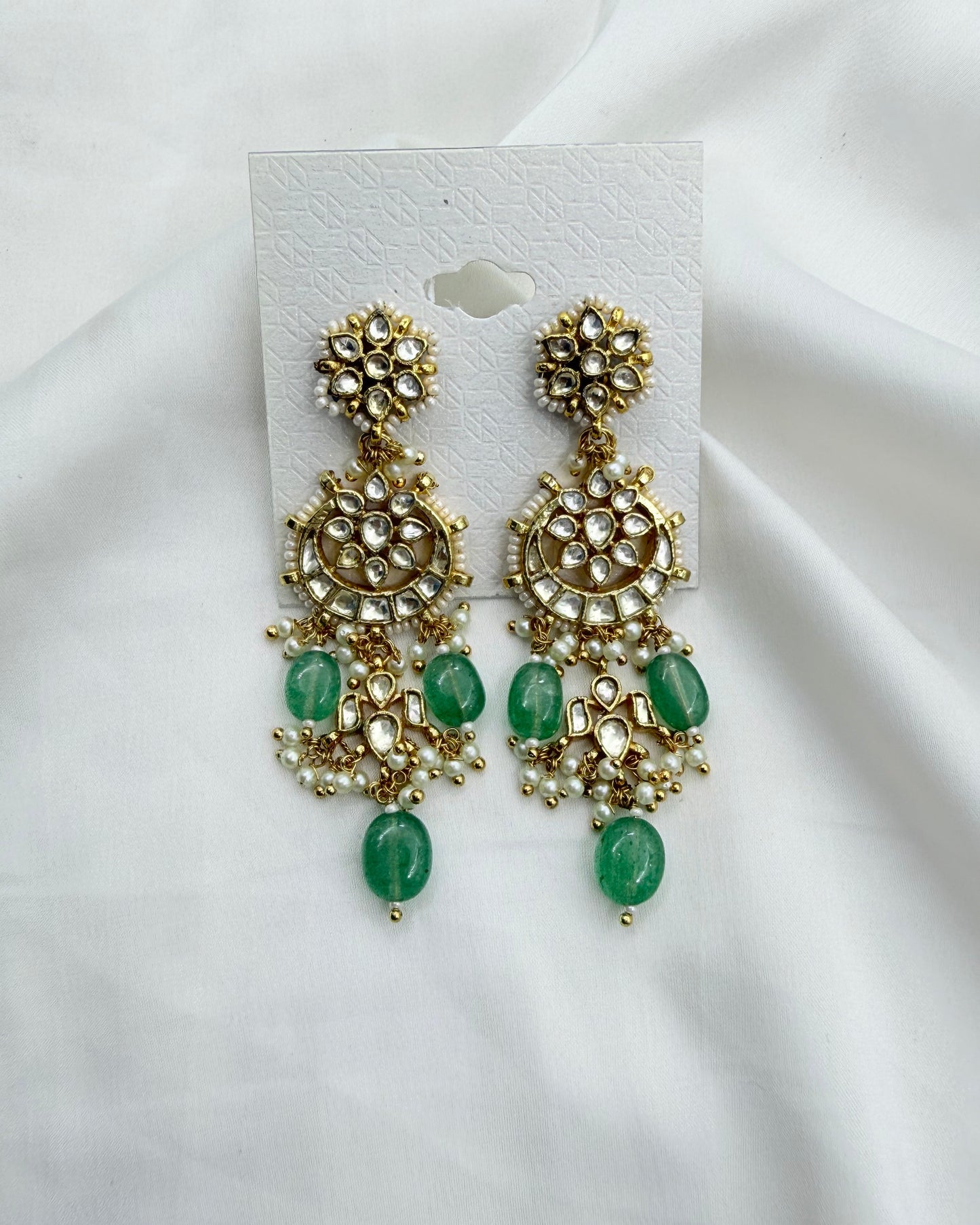 A pair of gold-toned chandelier earrings with green stone accents and clear rhinestone details, displayed on a white card against a white fabric background.