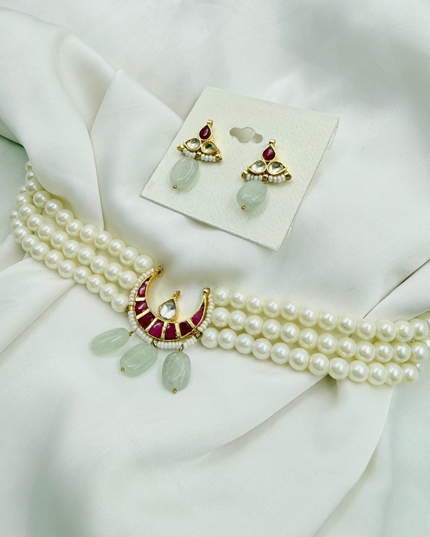 A traditional Indian jewelry set featuring a double-strand pearl necklace with a gold crescent pendant decorated with red stones and light green teardrop beads, alongside matching gold and pearl earrings on a display card, all arranged on a draped white fabric background.