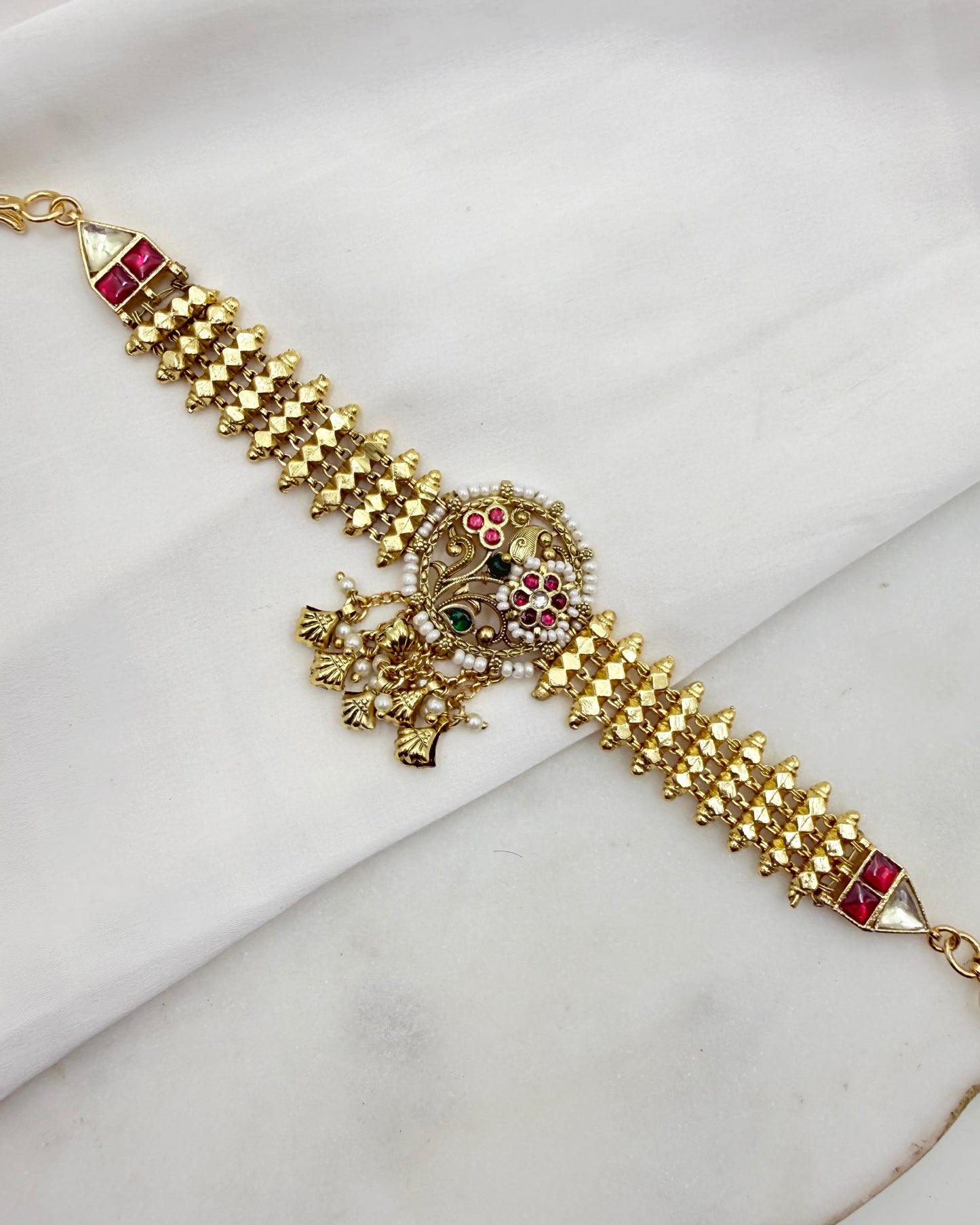 Gold-plated Indian Kundan bracelet with red and green stone embellishments, displayed diagonally on a white fabric background.
