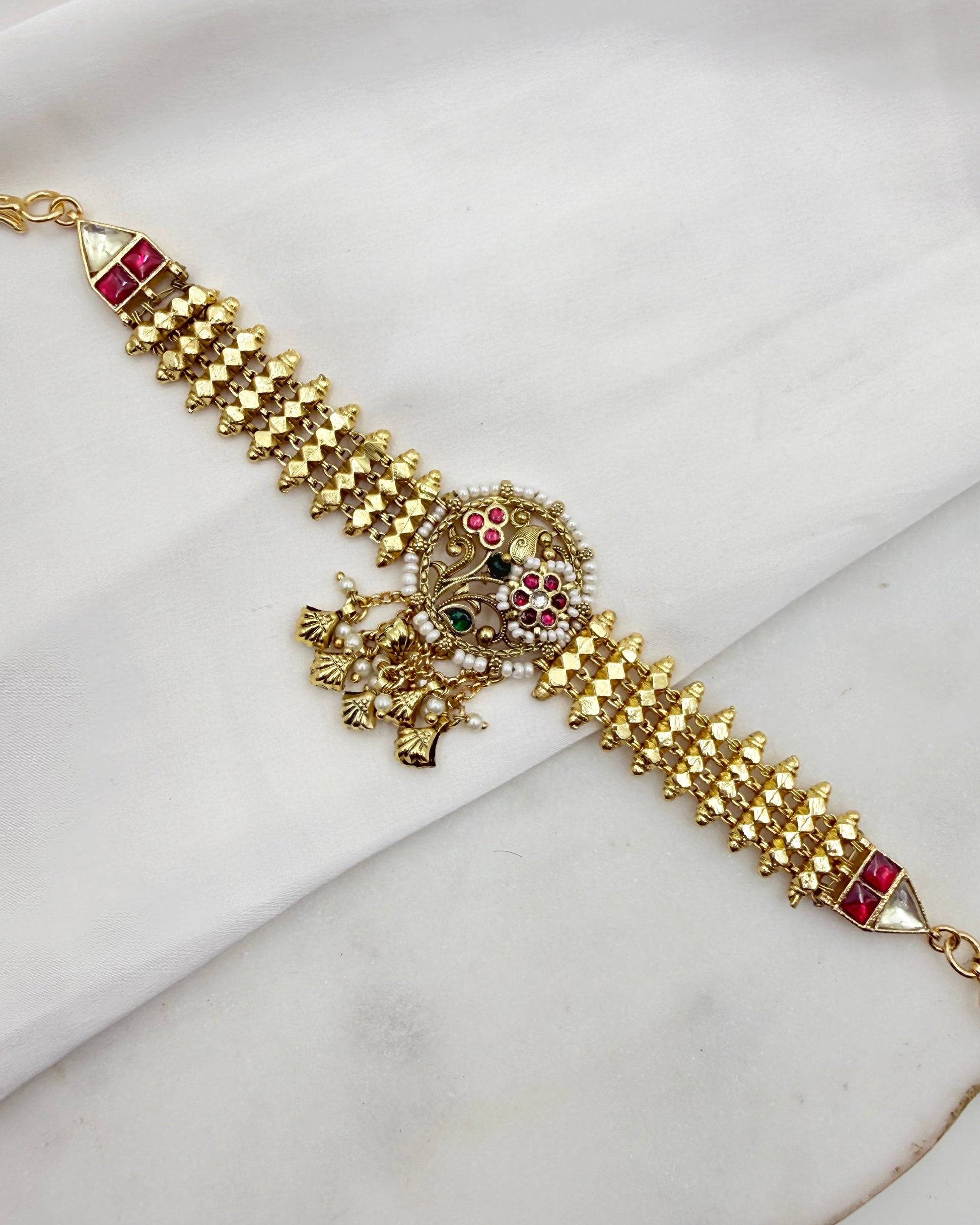 Gold-plated Indian Kundan bracelet with red and green stone embellishments, displayed diagonally on a white fabric background.