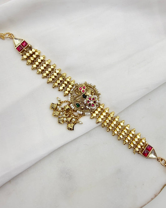 Gold-plated Indian Kundan bracelet with red and green stone embellishments, displayed diagonally on a white fabric background.