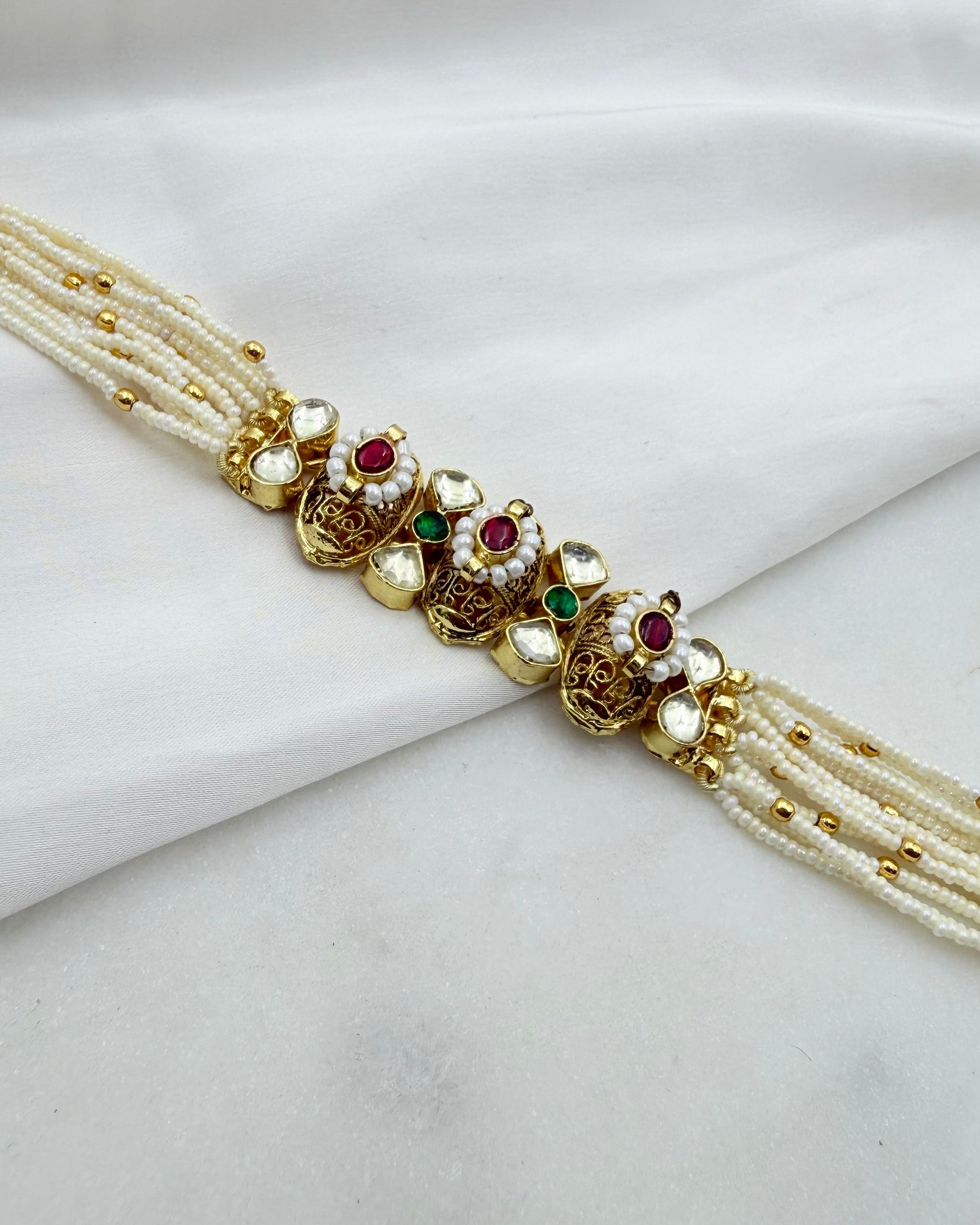 A traditional Indian gold-plated choker necklace with multiple strands of white pearl beads and decorative floral motifs set with white stones, red rubies, and green emeralds, displayed on a draped white fabric background.