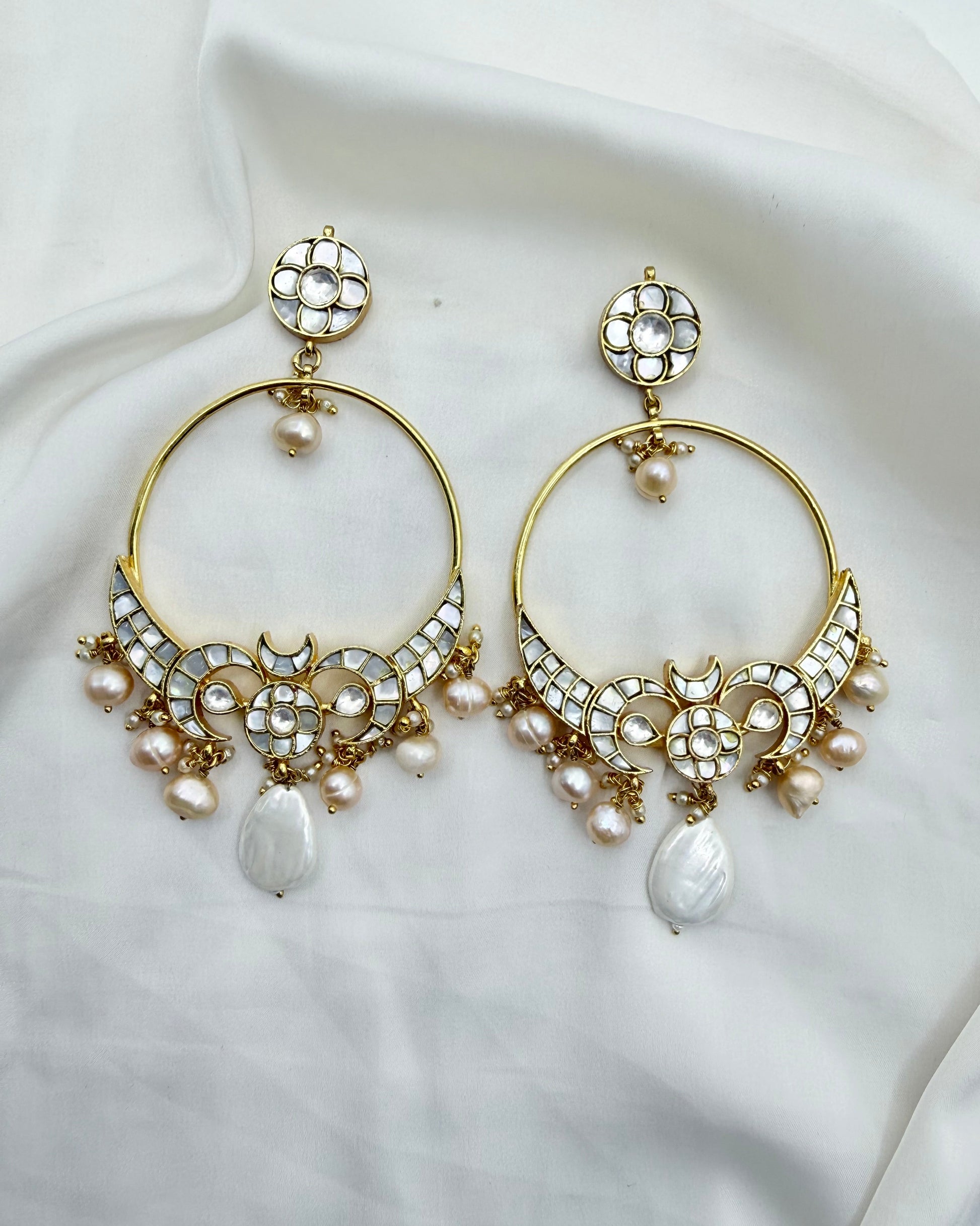 A pair of gold-plated hoop earrings adorned with white stones, small pearls, and teardrop pearl drops, displayed on a draped white fabric background.