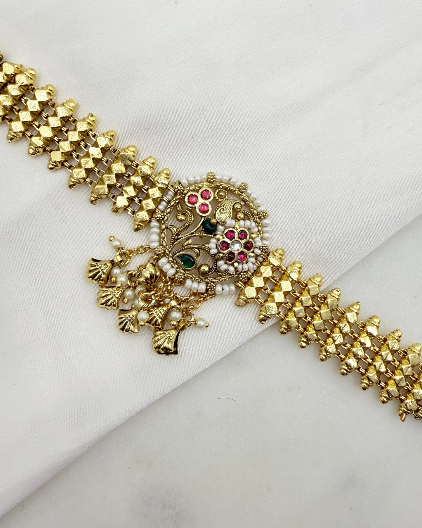 An ornate gold-toned traditional Indian jewelry piece, likely a choker or necklace, featuring a detailed central medallion with red, green, and white enamel accents, set against a draped white fabric background.