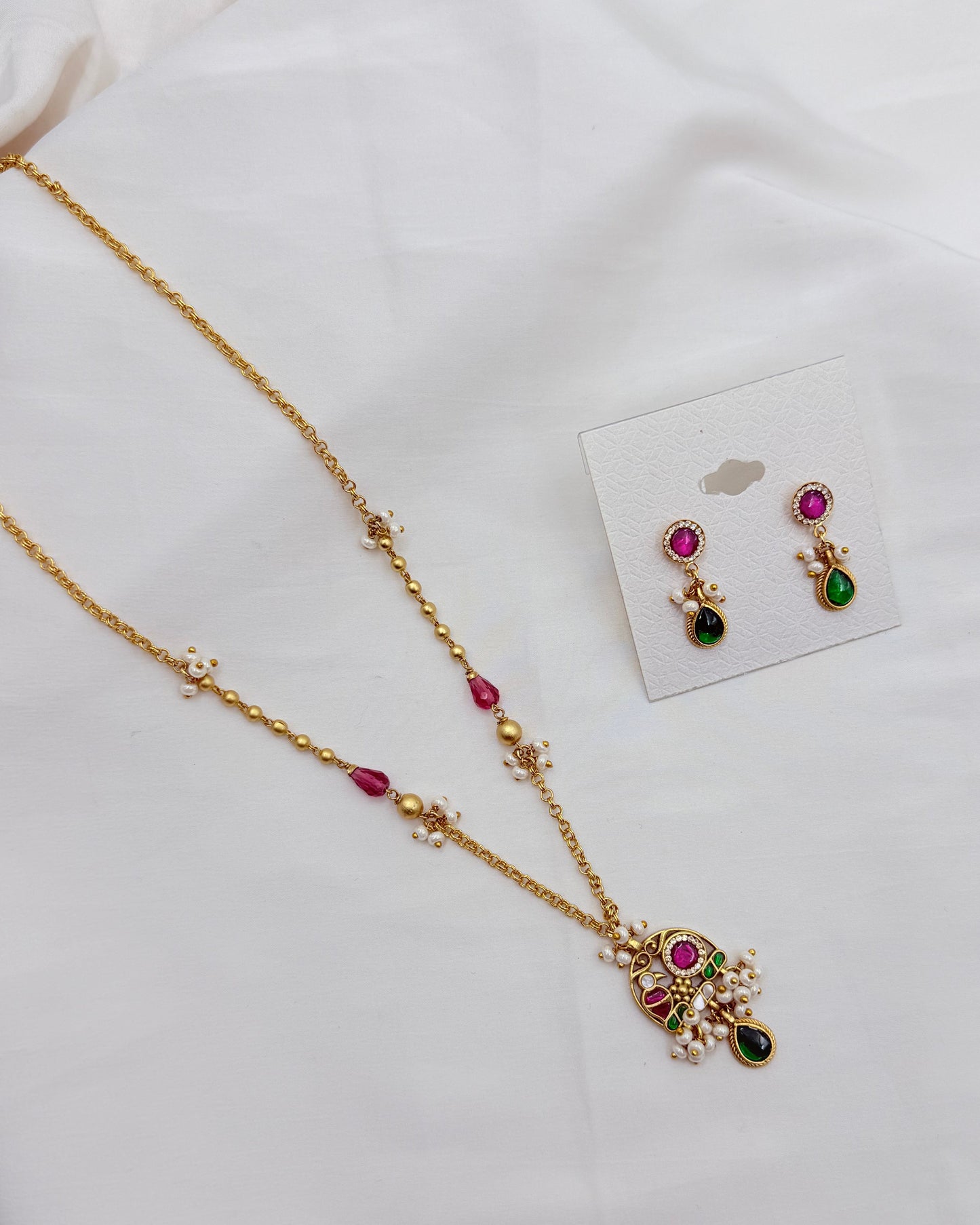 Gold-toned traditional jewelry set featuring a necklace and matching earrings with pink and green stones, displayed on a white fabric background with a small card holding the earrings.