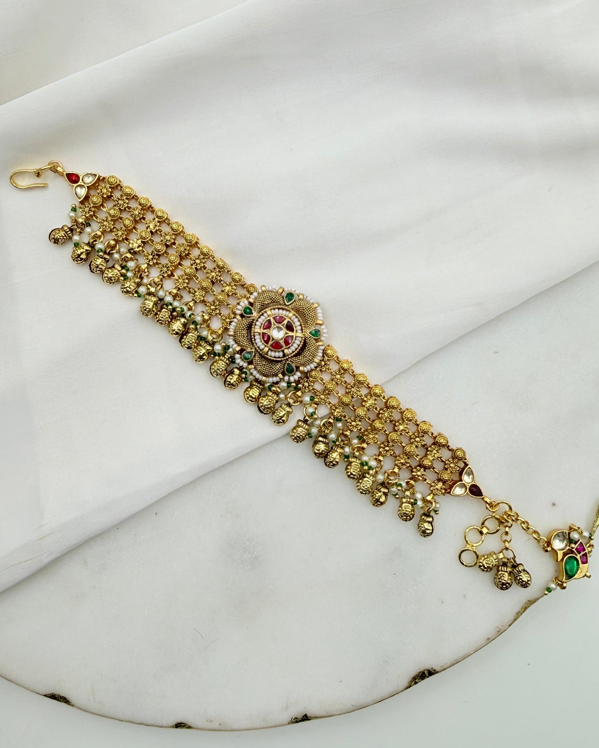 Gold-toned traditional Indian choker necklace with intricate beadwork, a central circular motif, and dangling bell details, displayed on a draped white fabric background.