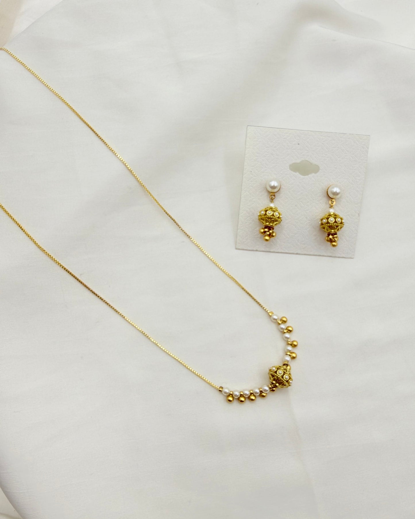 A gold necklace and matching drop earrings displayed on a white fabric background. The necklace has a delicate gold chain with a curved section of small gold beads leading to a larger textured gold ball pendant.
