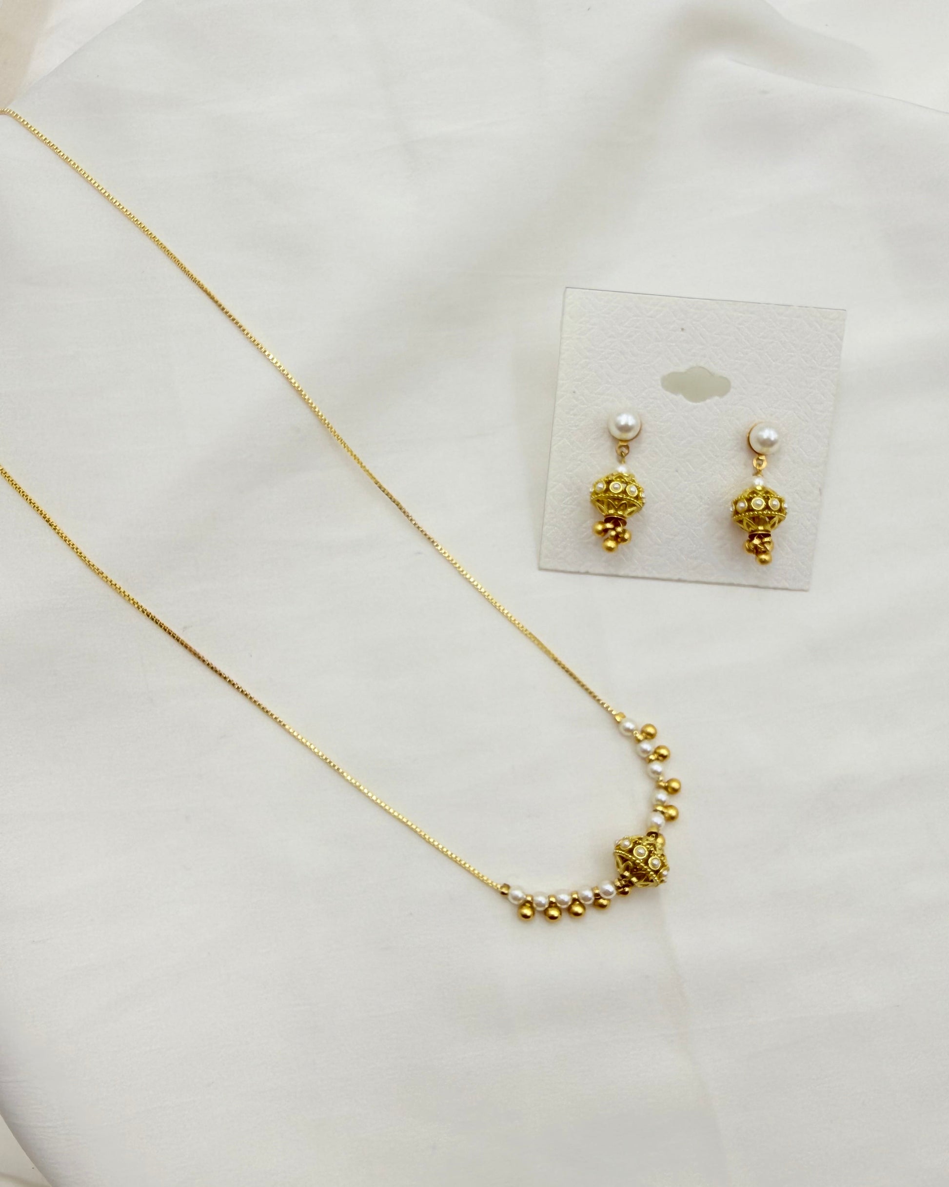 A gold necklace and matching drop earrings displayed on a white fabric background. The necklace has a delicate gold chain with a curved section of small gold beads leading to a larger textured gold ball pendant.