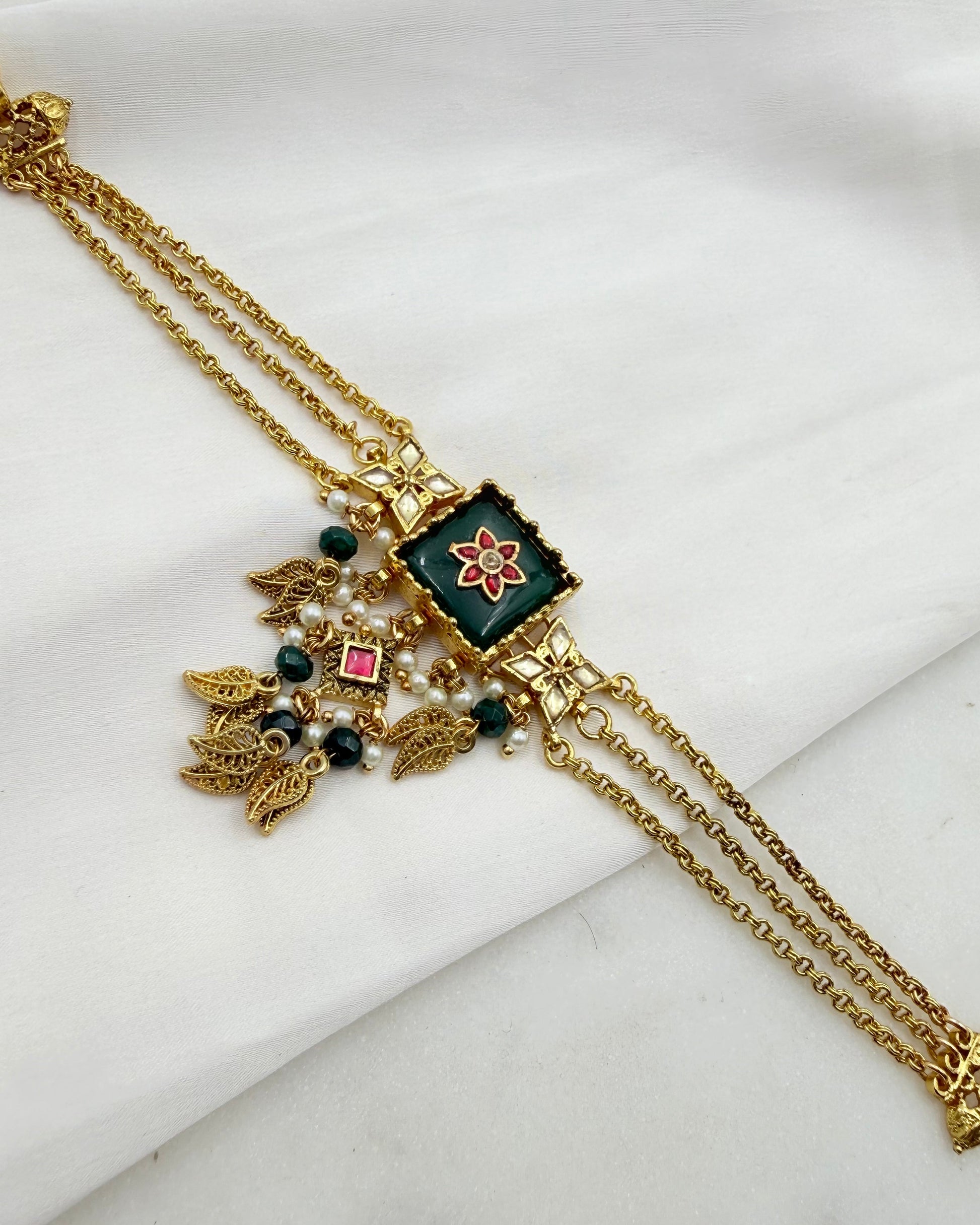 Gold-plated traditional Indian jewelry piece featuring a central square green enamel panel with a red floral motif, adorned with hanging gold leaf charms, small green beads, and a pink rhinestone, displayed on a white fabric background.