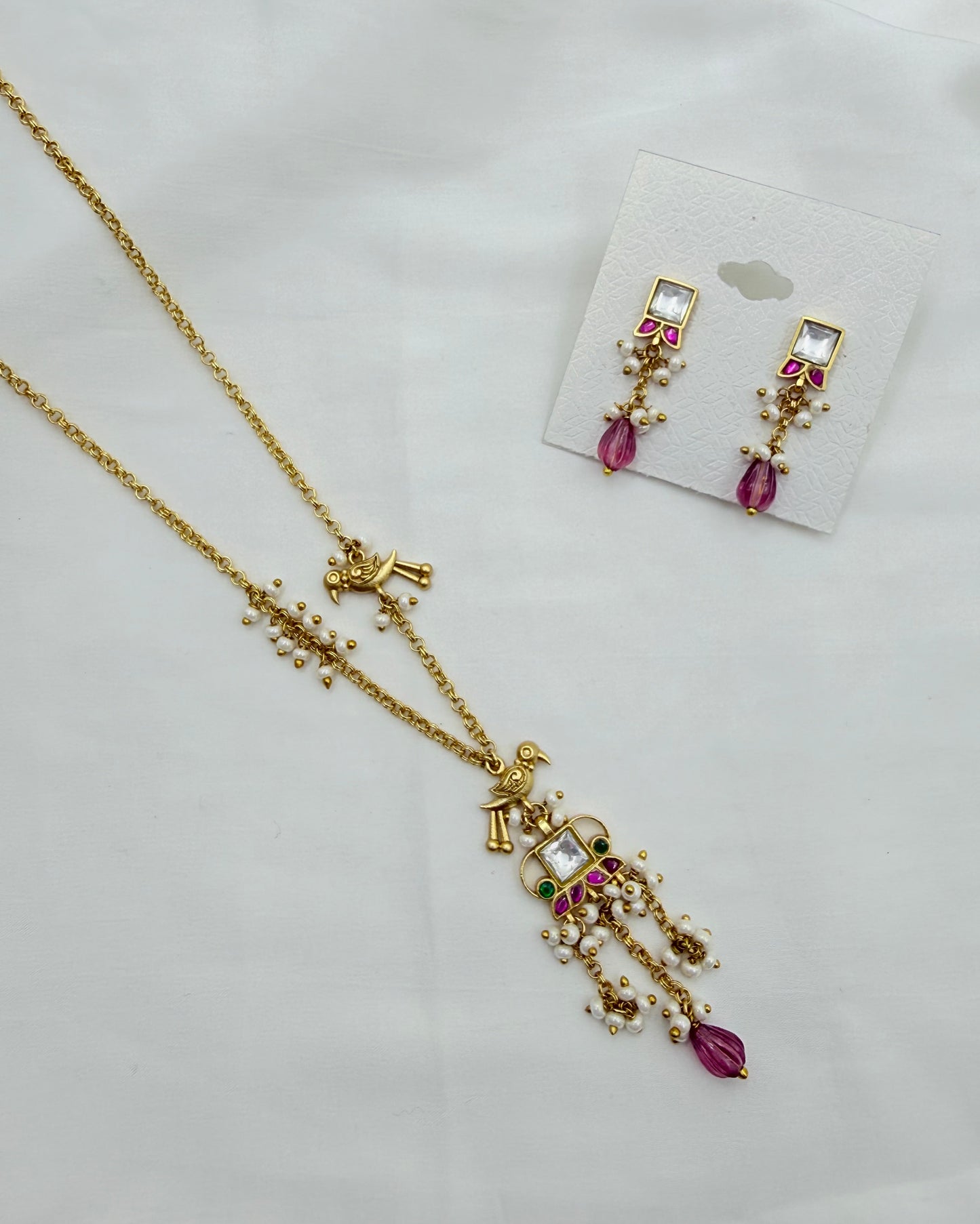 Gold-toned traditional jewelry set featuring a necklace and matching earrings with white stones and pink teardrop accents, displayed on a white fabric background.