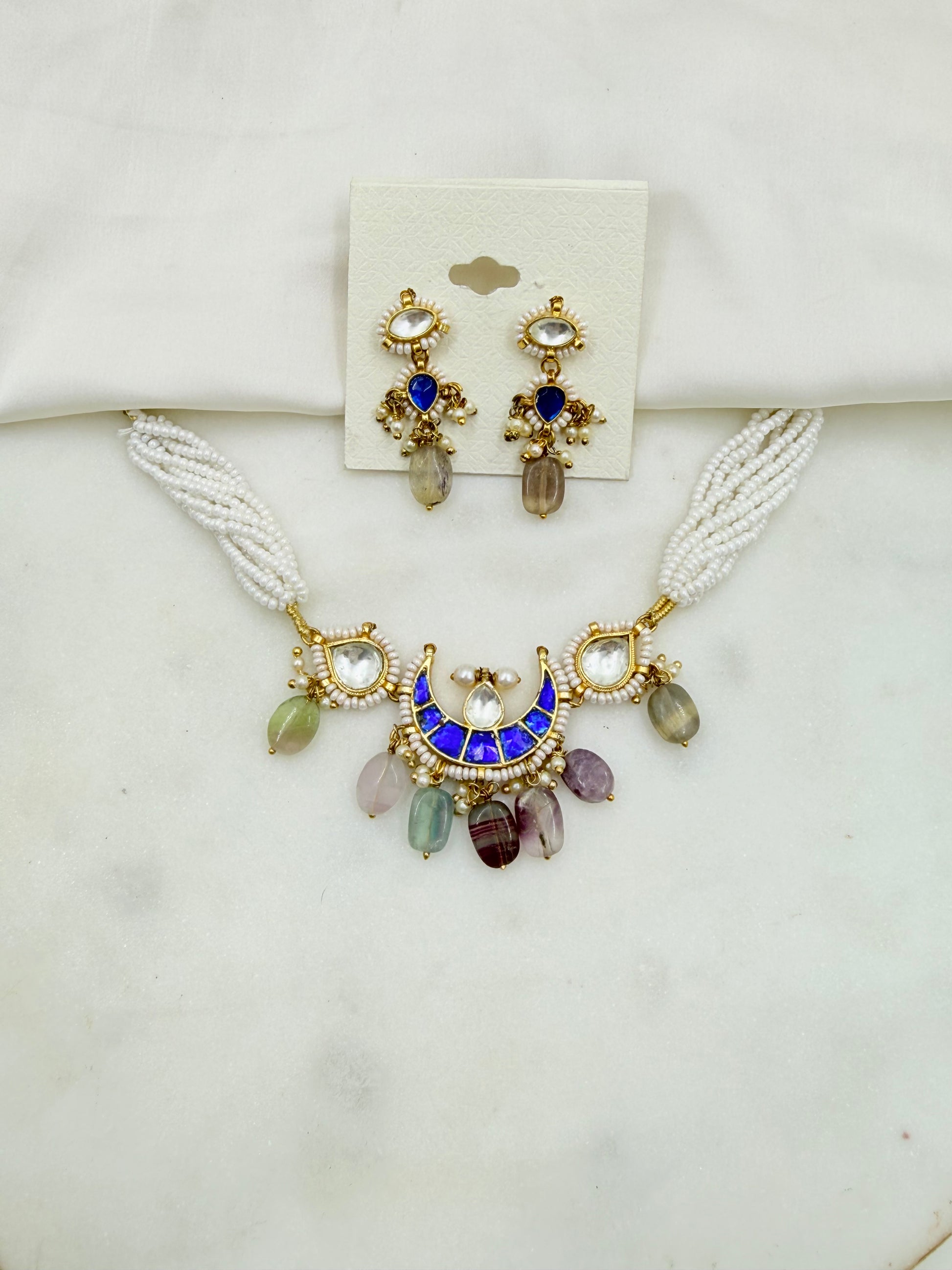 A gold-plated jewelry set featuring a necklace and matching earrings on a white display card, set against a light marble background. The necklace has a white beaded strap, a gold crescent moon pendant with a blue enamel center, and multicolored teardrop gemstones.