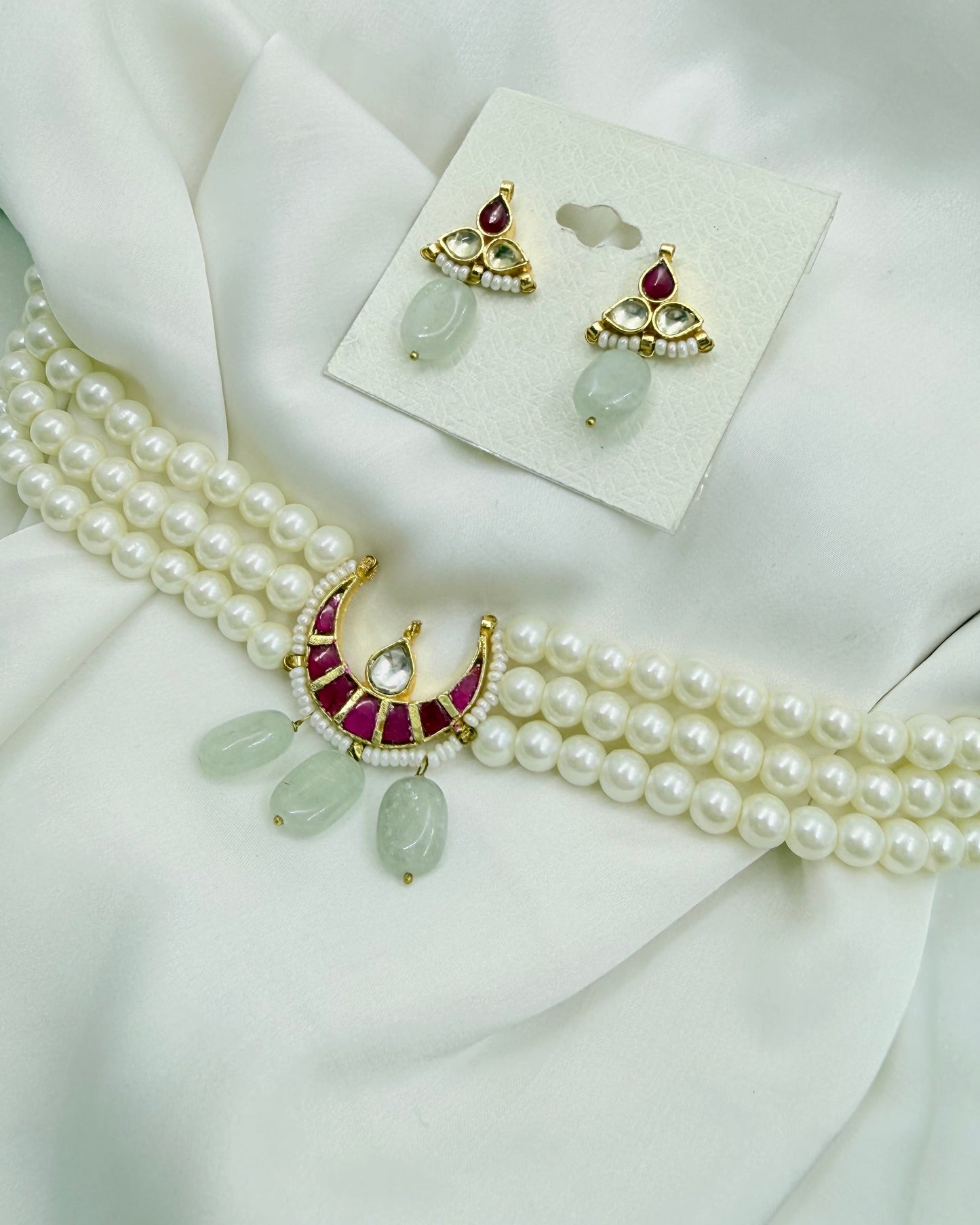 A gold and white pearl jewelry set featuring a multi-strand pearl necklace with a crescent moon pendant decorated with red stones and light green teardrop beads, alongside matching gold and pearl earrings on a white card, all arranged on a draped white satin fabric.