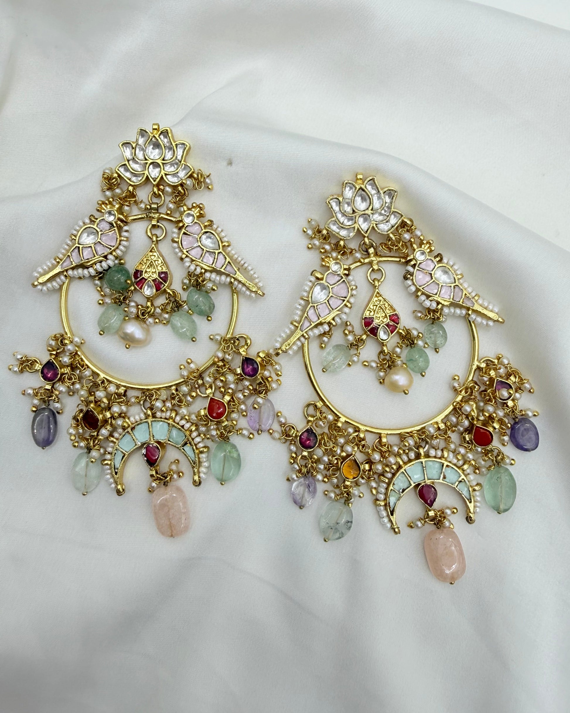 A pair of ornate gold-toned statement earrings with bird motifs, floral details, and dangling multicolored gemstones, displayed on a soft white fabric background.