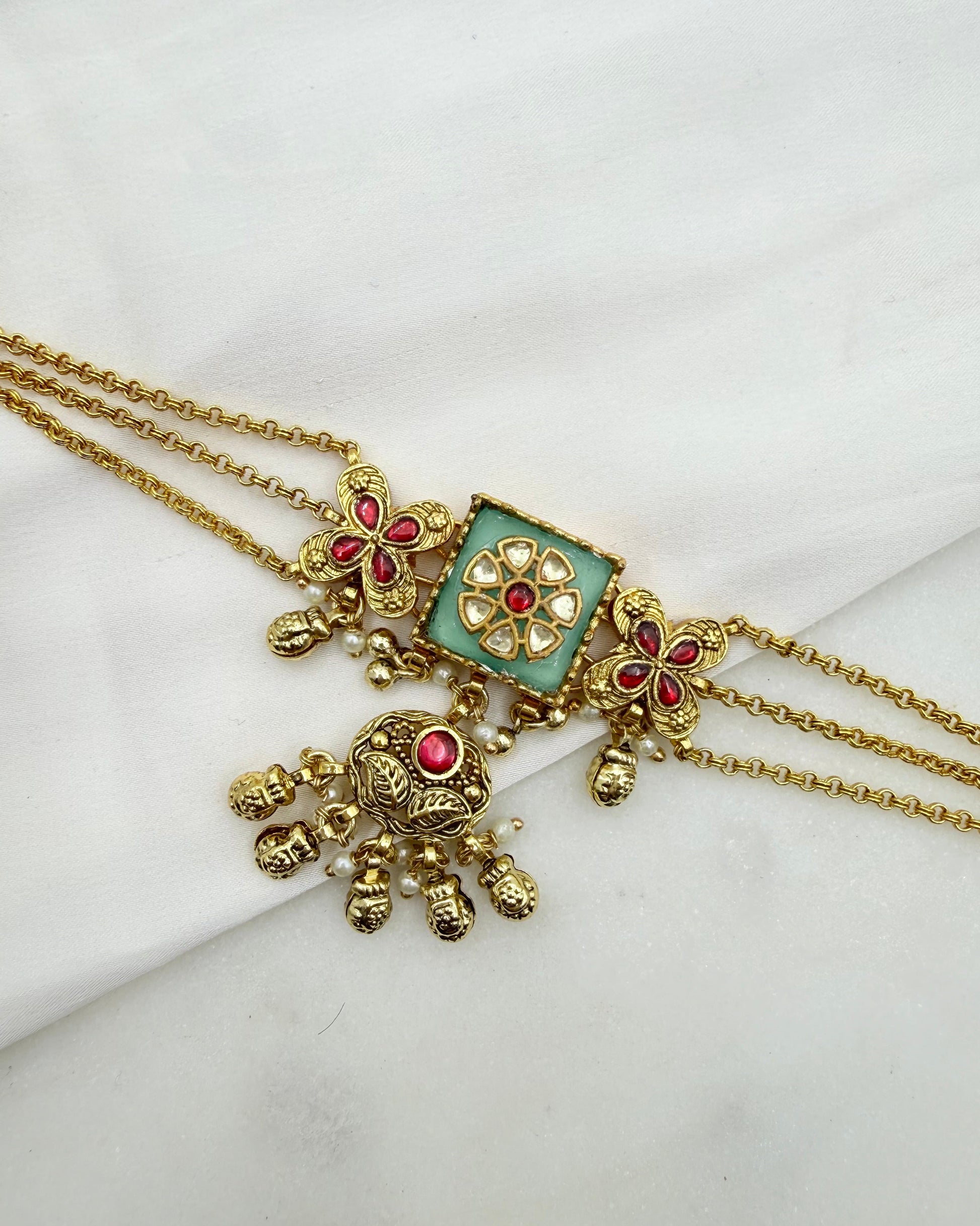 Gold-plated double-strand chain necklace with a square-shaped light green enamel center, decorated with gold floral details and red stones, resting on a white fabric background.
