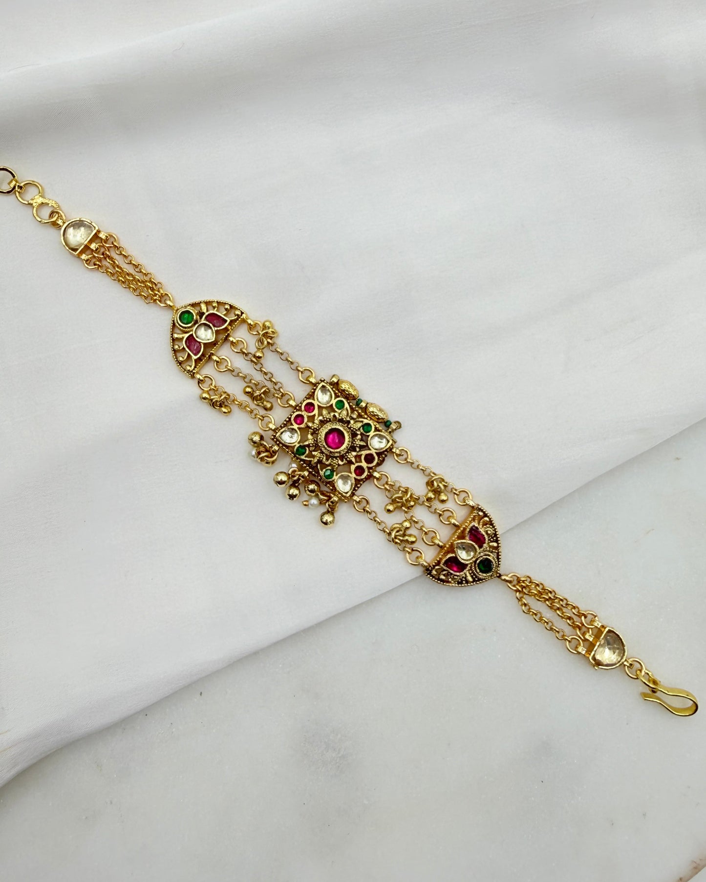 Gold-toned traditional Indian Kundan bracelet with intricate filigree work, red and green stone embellishments, and hanging chain details, displayed on a white fabric background.