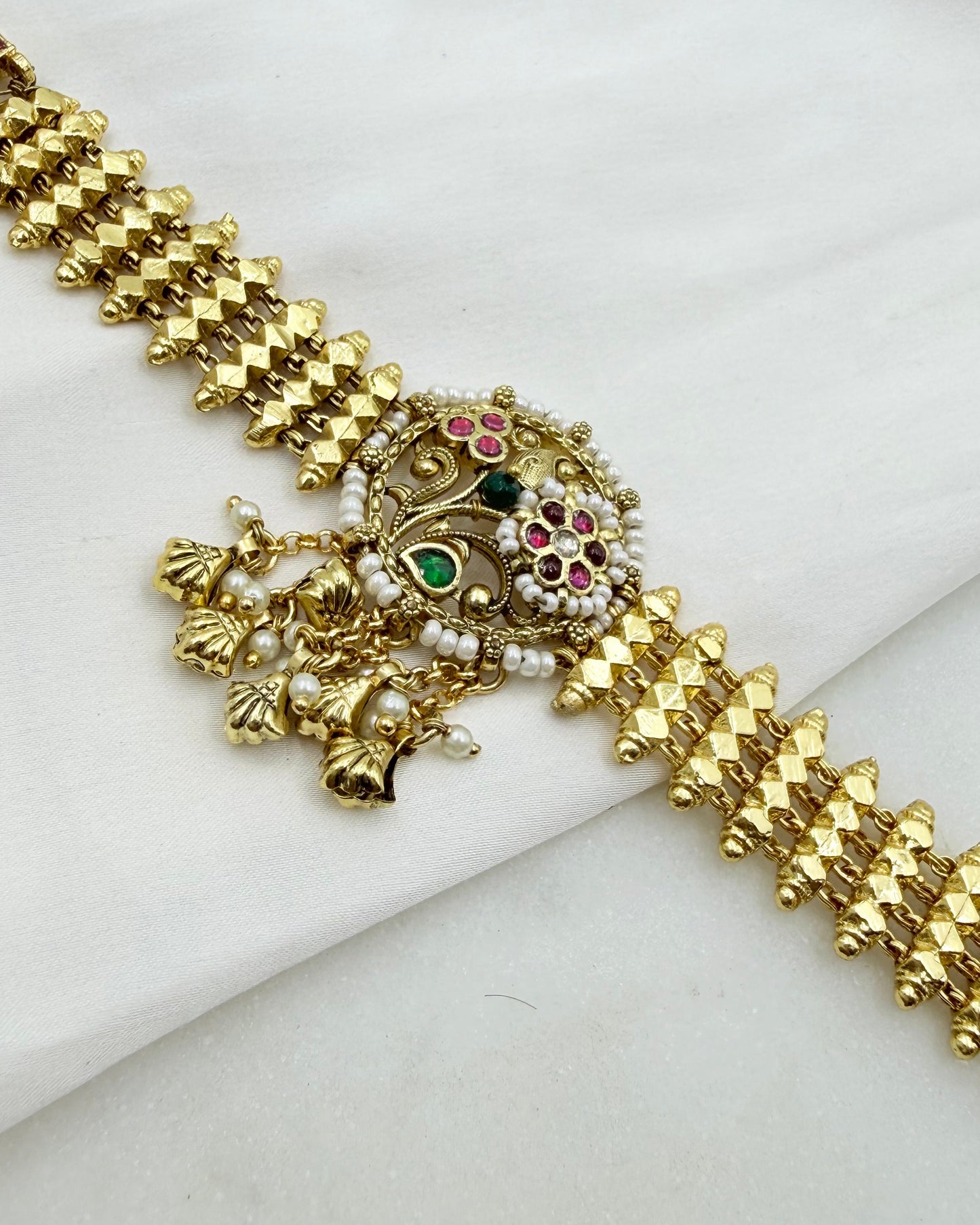 Gold-plated traditional Indian choker bracelet with a central ornate pendant featuring green, pink, and white stones, displayed on a white fabric background.