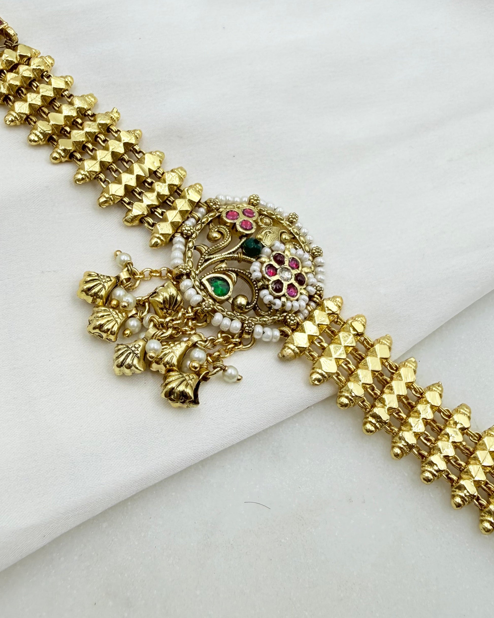 Gold-plated traditional Indian choker bracelet with a central ornate pendant featuring green, pink, and white stones, displayed on a white fabric background.