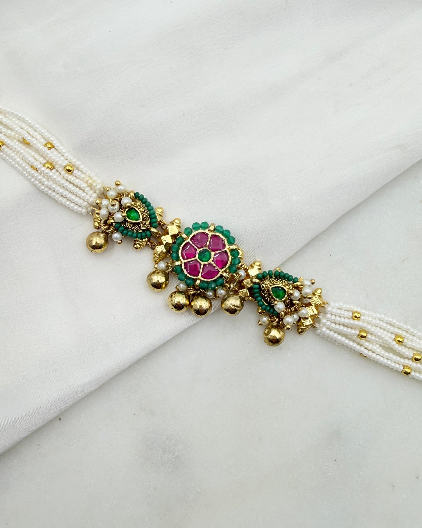 A traditional Indian multi-strand pearl bracelet with gold-plated borders and decorative floral motifs in pink, green, and white, displayed on a white fabric background.
