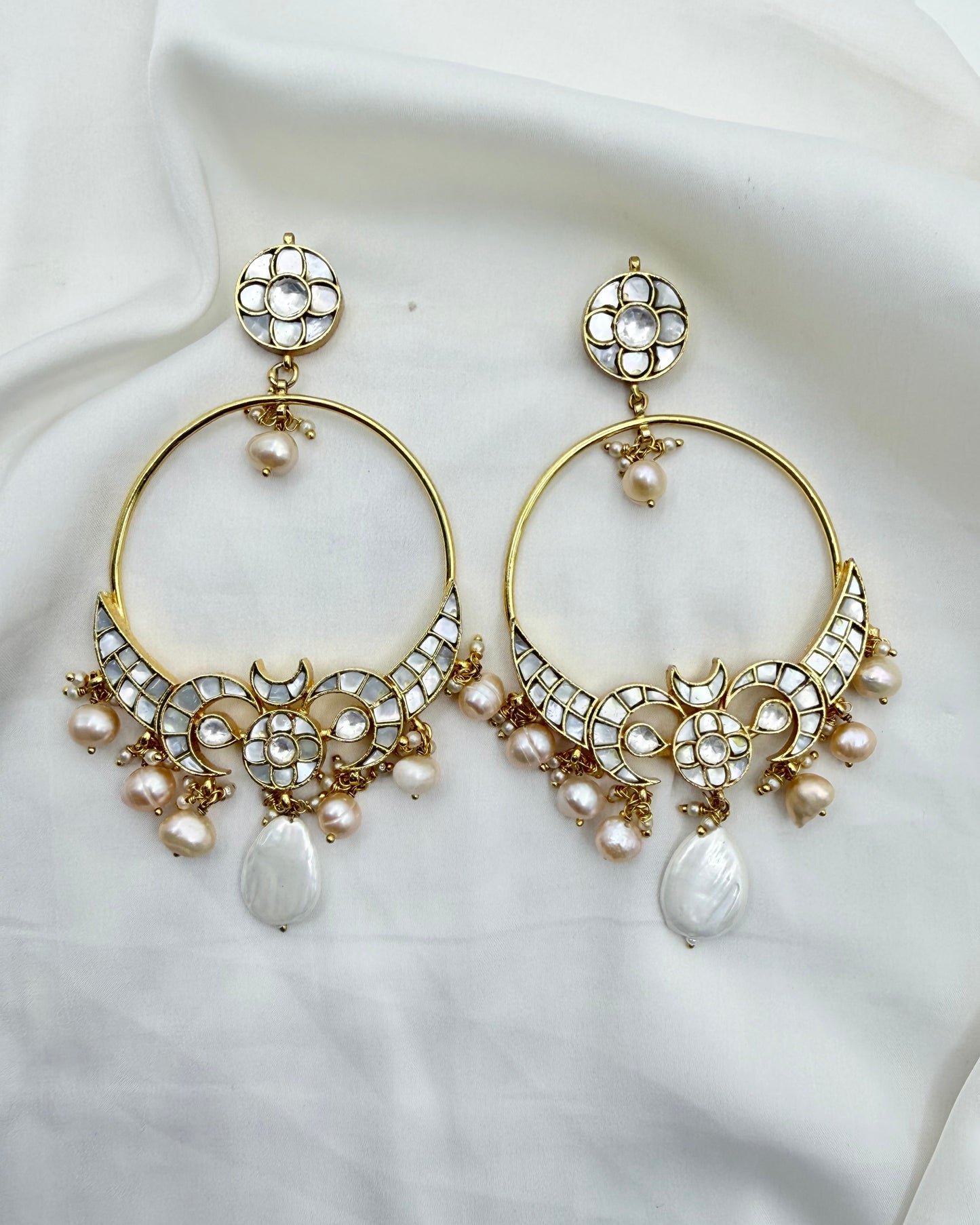 A pair of gold-toned huggie hoop earrings decorated with white stones, small pearls, and teardrop pearl drops, displayed on a draped white fabric background.