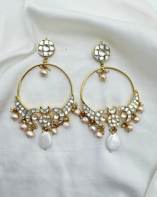 A pair of gold-toned huggie hoop earrings decorated with white stones, small pearls, and teardrop pearl drops, displayed on a draped white fabric background.