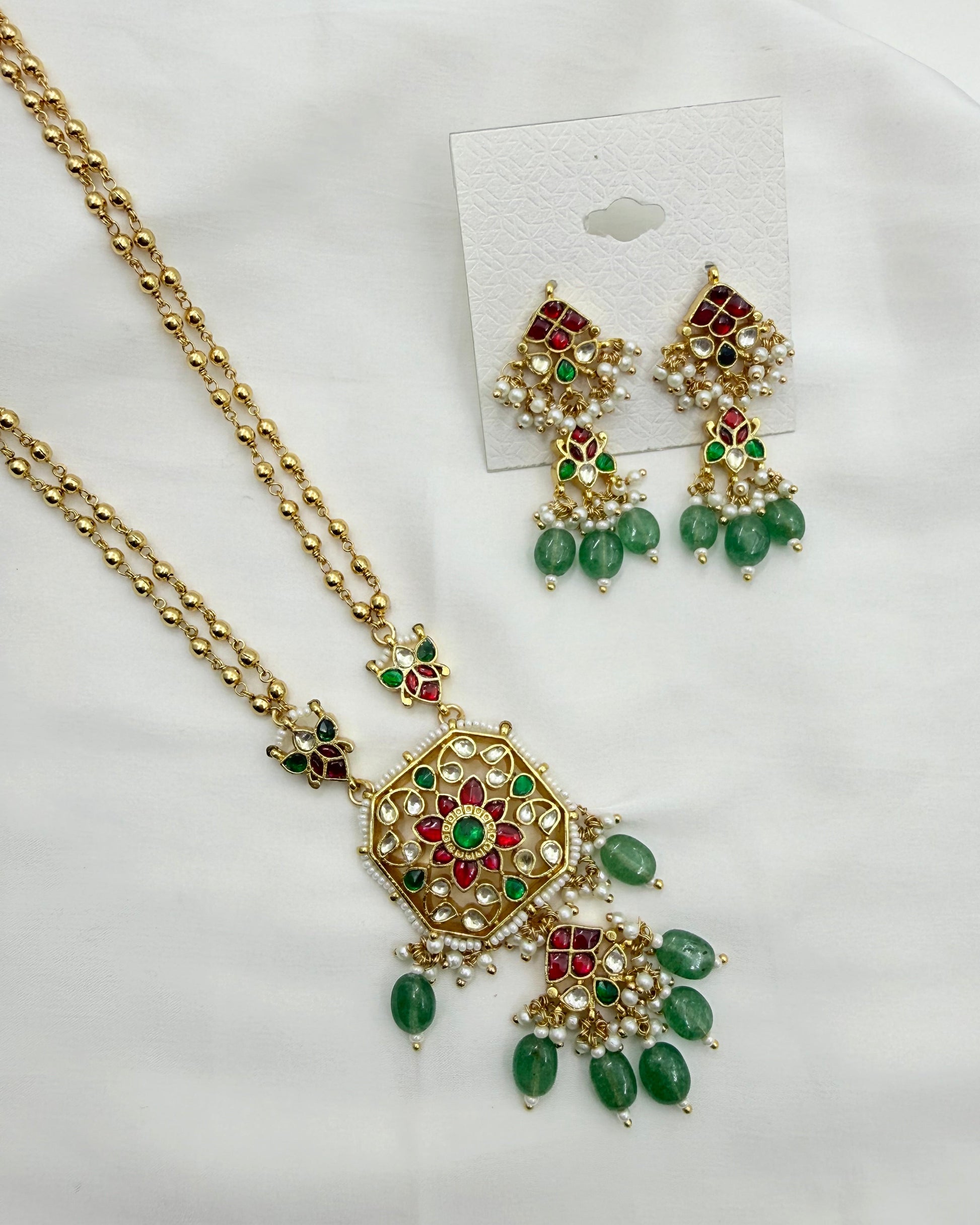 Gold-toned Indian jewelry set featuring a necklace and matching earrings with green stone accents and red floral details, displayed on a white fabric background.