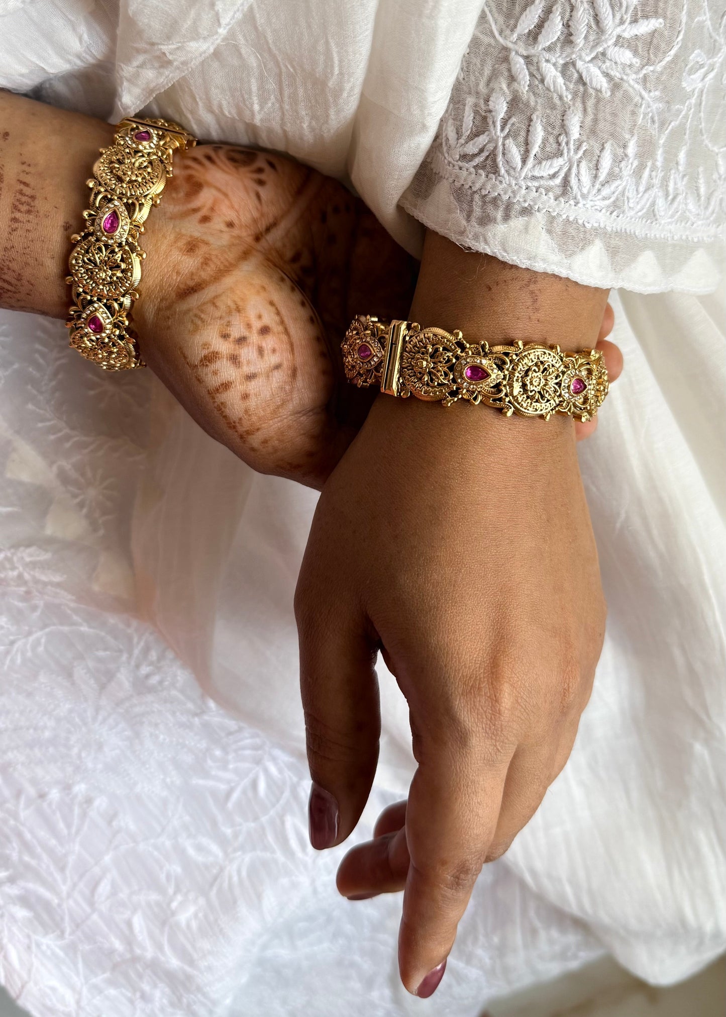 Close-up of hands wearing gold bracelets with pink stones on a white lace fabric background.